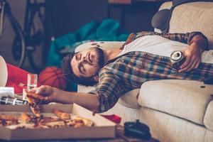 esso ha stato un' lungo notte. giovane bello uomo passato su su divano con Pizza fetta e birra può nel il suo mano nel disordinato camera dopo festa foto