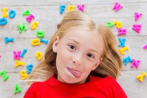 sfacciato poco ragazza. superiore Visualizza di carino poco ragazza smorfie mentre dire bugie su il pavimento con plastica colorato lettere posa in giro sua foto