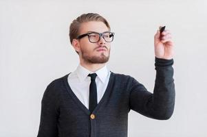 scrittura su trasparente pulire tavola. bello giovane uomo nel bicchieri conservazione braccia attraversato e mentre in piedi contro grigio sfondo foto