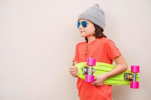 fiducioso pattinatore. lato Visualizza di fiducioso poco ragazzo nel cappello Tenere colorato skateboard mentre in piedi contro grigio sfondo foto