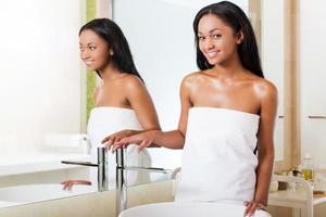 fresco e pulito bellezza. bellissimo giovane africano donna avvolto nel un' asciugamano guardare a telecamera e sorridente mentre in piedi contro un' specchio nel bagno foto