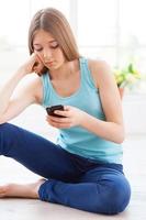 in attesa per il suo chiamata. depresso adolescenziale ragazza Tenere mobile Telefono e guardare a esso mentre seduta su il pavimento a sua appartamento foto