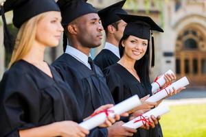 contento laureato. quattro Università laureati in piedi nel un' riga e Tenere loro diplomi mentre uno donna guardare a telecamera e sorridente foto