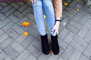 un' bellissimo sottile ragazza, un' donna raddrizza, tocchi sua gambe, jeans, pantaloni e stivali, scarpe seduta su un' parco panca. il sfondo foto