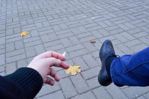 un' uomo detiene un' sigaretta nel il suo mani, dita seduta nel il parco e fumo con il suo gamba nel un' scarpa contro il sfondo di pavimentazione lastre con autunno le foglie. il sfondo foto