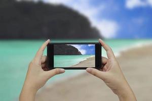 mano Tenere smartphone con bianca vuoto schermo al di sopra di sfocato sulla spiaggia montagne sfondo foto