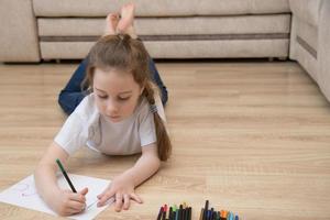 carino poco ragazza nel jeans bugie su il pavimento a casa e disegna con colorato matite, il concetto di il sviluppo di un' bambino in età prescolare, Hobby foto