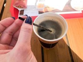 un' donna detiene con un' bellissimo manicure su sua dita un' tubo nel un' tazza di veloce nero caldo forte naturale caffè a partire dal veloce cibo nel un' carta bicchiere foto