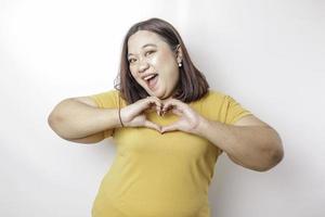 un attraente giovane asiatico grande dimensione donna indossare un' giallo maglietta si sente contento e un' romantico forme cuore gesto esprime tenero sentimenti foto