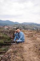 elegante giovane uomo su il ferrovia ponte nel vorokta, Carpazi contro il fondale di un' montagna paesaggio. turisti seduta nel il montagne vicino il ferrovia foto