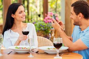 Sorridi per il telecamera bello giovane uomo assunzione immagine di sua bellissimo fidanzata con inteligente Telefono mentre rilassante nel all'aperto ristorante insieme foto