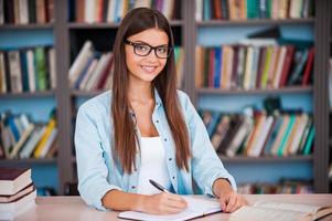 così contento per essere un' alunno bellissimo giovane donna scrittura qualcosa nel sua Nota tampone e guardare a telecamera mentre seduta a il scrivania nel il biblioteca foto