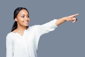 appena Guarda a quello attraente giovane africano donna puntamento lontano e sorridente mentre in piedi contro grigio sfondo foto