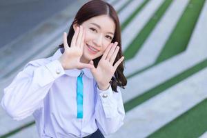 bellissimo alto scuola asiatico alunno ragazza nel il scuola uniforme sta e sorrisi felicemente con bretelle su sua denti mentre lei mettere sua mano su viso con fiducia mentre lei sembra a il telecamera foto