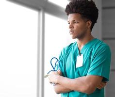 premuroso giovane medico afroamericano in piedi vicino alla finestra distogliendo lo sguardo braccia incrociate. il medico maschio del ritratto tiene lo stetoscopio seriamente pensando, meditando. stile di vita professionale di occupazione medica. foto