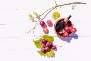 cosmetico olio, siero basato su uva seme olio nel un' bicchiere fiala con un' pipetta su un' di legno ciotola con un' mazzo di uva. superiore Visualizza. bianca di legno sfondo. foto