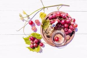 cosmetico olio, siero basato su uva seme olio nel un' bicchiere fiala con un' pipetta su un' di legno vassoio con un' mazzo di uva. superiore Visualizza. bianca di legno sfondo. foto