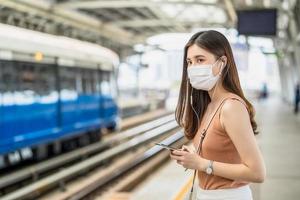 Passeggero di giovane donna asiatica che indossa maschera chirurgica e ascolta musica tramite telefono cellulare intelligente in treno della metropolitana quando si viaggia in una grande città allo scoppio di covid19, infezione e concetto di pandemia