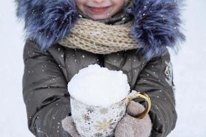 un' ragazza nel caldo inverno all'aperto Abiti detiene un' boccale con neve nel sua mani - freddo tempo metereologico condizioni, stagionalità. Natale e nuovo anno foto