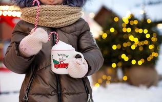 ragazza con boccale con neve, caramella canna e iscrizione allegro e luminosa nel sua mani all'aperto nel caldo Abiti nel inverno a festivo mercato. Fata luci ghirlande decorato neve cittadina per nuovo anno. Natale foto