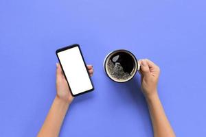 femmina mani Tenere nero mobile Telefono con vuoto bianca schermo e boccale di caffè. modello Immagine con copia spazio. superiore Visualizza su viola sfondo, piatto posare foto