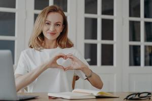 amichevole grato femmina Spettacoli cuore mano gesto seduta a il computer portatile su posto di lavoro. positivo risposta foto