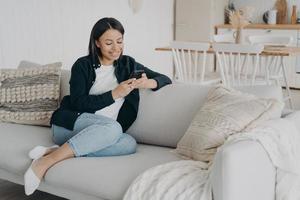 sorridente femmina usi Telefono app, chat nel sociale reti, shopping in linea, si siede su divano a casa foto