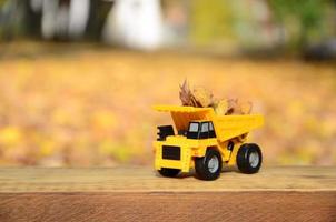un' piccolo giocattolo giallo camion è caricato con giallo caduto le foglie. il auto sta su un' di legno superficie contro un' sfondo di un' sfocato autunno parco. pulizia e rimozione di caduto le foglie. di stagione lavori foto