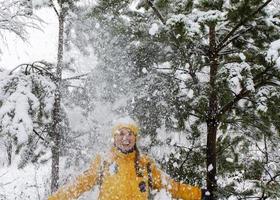 giovane ridendo contento caucasico donna nel giallo giacca sotto caduta neve a partire dal pino rami nel inverno foresta. positivo emozioni, a piedi nel nevoso tempo metereologico foto