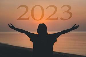 2023 contento nuovo anno concetto, salutare donna sollevato mano Tenere 2023 personaggio testo a Alba su il spiaggia. foto