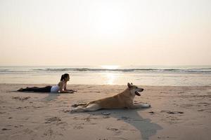 giovane salutare donna e cane fare verso l'alto cane yoga posizione su il spiaggia. foto