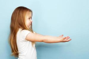 poco bionda ragazza con lungo capelli teso sua braccia per il lato. bambino Tenere Aperto palma vuoto mano su blu sfondo foto