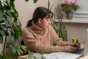 bellissimo adolescente ragazza fioraio casa giardiniere con casa fiore su tavolo utilizzando in linea video Istruzioni su il computer portatile a casa su tavolo. giovane femmina libero professionista Lavorando su un' il computer portatile foto