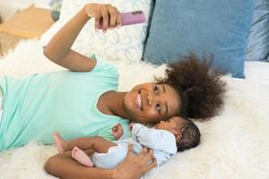 giovane madre africano americano e neonato bambino fabbricazione video chiamata e sorridente salutare. contento Sambuco sorella assunzione autoscatto con poco fratello infantile dire bugie su letto guardare a mobile Telefono sorridente spensierato. foto