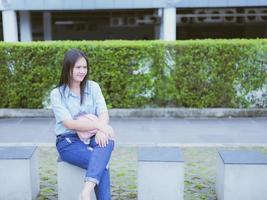 prendere un' ritratto di un asiatico donna con nero capelli nel un' blu camicia e jeans. sedersi su il nono nel il parco e ha un' sfondo di edifici e cespugli. foto