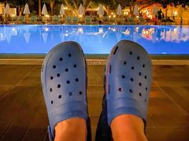 piedi nel gomma da cancellare blu Flip flop, scarpe nel davanti di un' piscina di acqua nel un tutti compreso Hotel a notte nel un' caldo tropicale orientale Paradiso Sud ricorrere foto