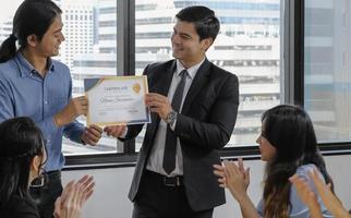 giovane caucasico manager nel completo da uomo dando finto su certificato per asiatico personale con contento sorridente viso mentre altri Applaudire per congratulazione nel ufficio incontro camera. selettivo messa a fuoco. foto