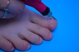 il processi di fare un' pedicure su il dita dei piedi di il chiodo in lavorazione di il chiodo con il Aiuto di un' speciale macchina su un' blu sfondo foto