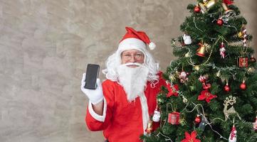 Santa Claus festeggiare Natale nel felicità e eccitazione mentre Tenere mobile Telefono accanto il completamente decorato Natale albero per vendita promozione e in linea shopping uso foto