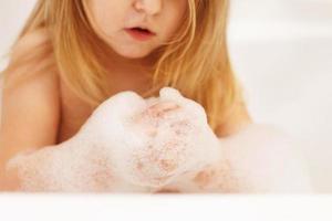 divertente poco bambino ragazza con biondo capelli giocando con schiuma nel un' bagno vasca. ragazza prende un' bagno foto