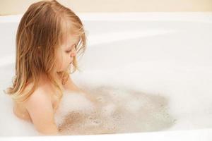 divertente poco bambino ragazza con biondo capelli giocando con schiuma nel un' bagno vasca. ragazza prende un' bagno foto