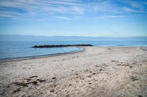 sabbioso spiaggia su il costa di Danimarca. pietra frangiflutti nel il baia, luce del sole mentre a piedi foto