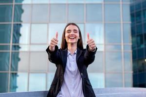 ritratto di un' riuscito attività commerciale donna sorridente mostrando pollici su. bellissimo giovane femmina esecutivo nel un urbano ambientazione foto