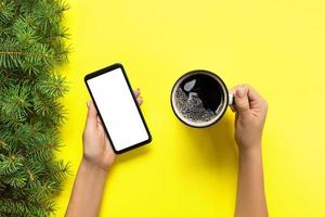 femmina mani Tenere nero mobile Telefono con vuoto bianca schermo e boccale di caffè. modello Immagine con copia spazio. superiore Visualizza su giallo sfondo, piatto posare foto