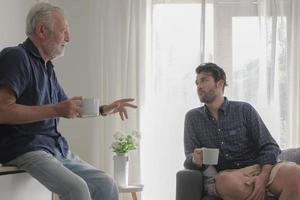 vecchio caucasico padre potabile caffè e rilassare parlando con il suo figlio durante vacanza nel accogliente vivente camera Casa sfondo. condivisione famiglia storia e autentico stile di vita concetto. foto