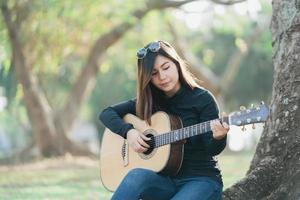 asiatico femmina musicisti indossare un' nero maniche lunghe camicia e occhiali da sole su sua testa. cantando e giocando acustico chitarra sotto un' albero nel il parco nel il mattina. foto