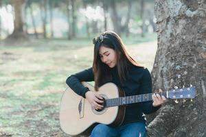 asiatico femmina musicisti indossare un' nero maniche lunghe camicia e occhiali da sole su sua testa. cantando e giocando acustico chitarra sotto un' albero nel il parco nel il mattina. foto