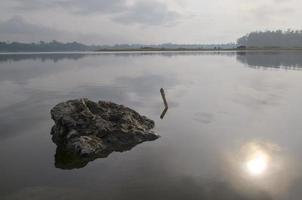 davanti Visualizza di un' grande roccia nel karangkate lago Indonesia con calma acqua condizioni a Alba con un' montagne sfondo e un' nebbioso cielo foto