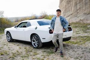 bello uomo nel jeans giacca e berretto a piedi vicino il suo bianca muscolo auto nel carriera. foto