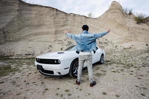 indietro di uomo nel jeans giacca e berretto è in piedi e mani per il lati, vicino il suo bianca muscolo auto nel carriera. los angeles. foto